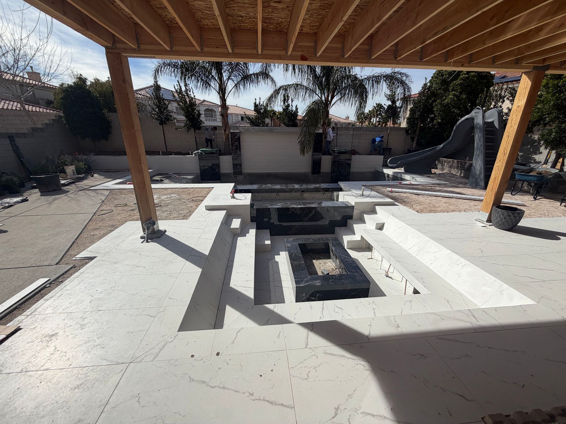 A sunken patio with a black fire pit and seating area under a wooden pergola, viewed from a light-colored concrete deck.