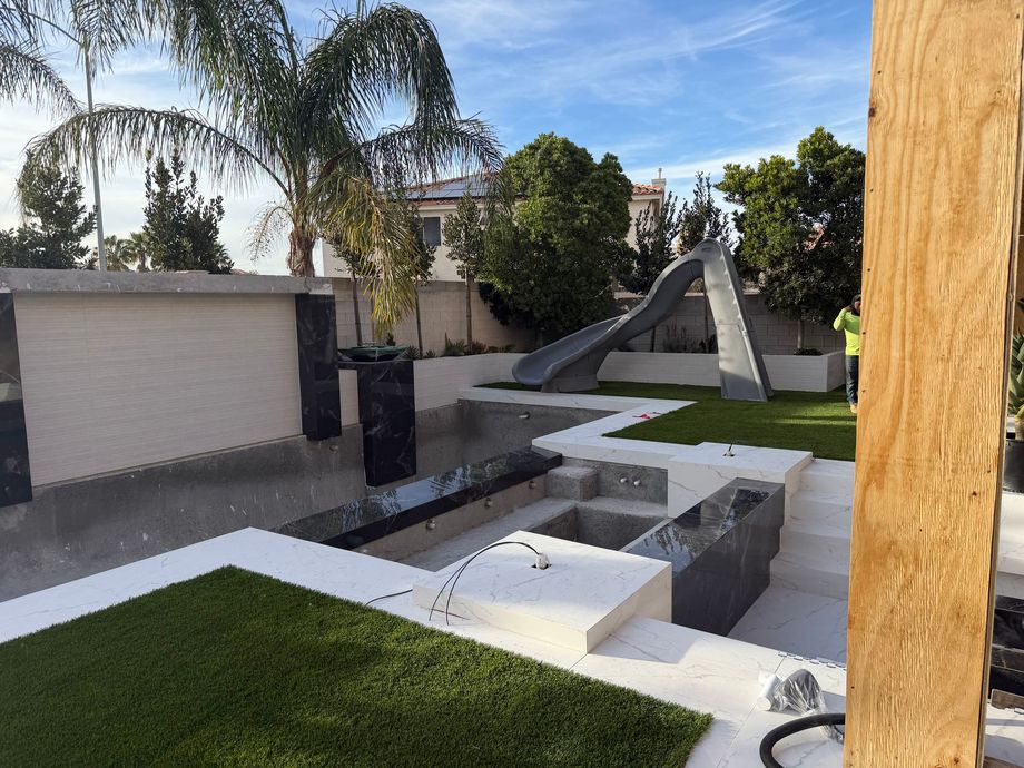 A backyard pool under construction with white tiered concrete levels, a gray slide, and patches of new artificial grass.