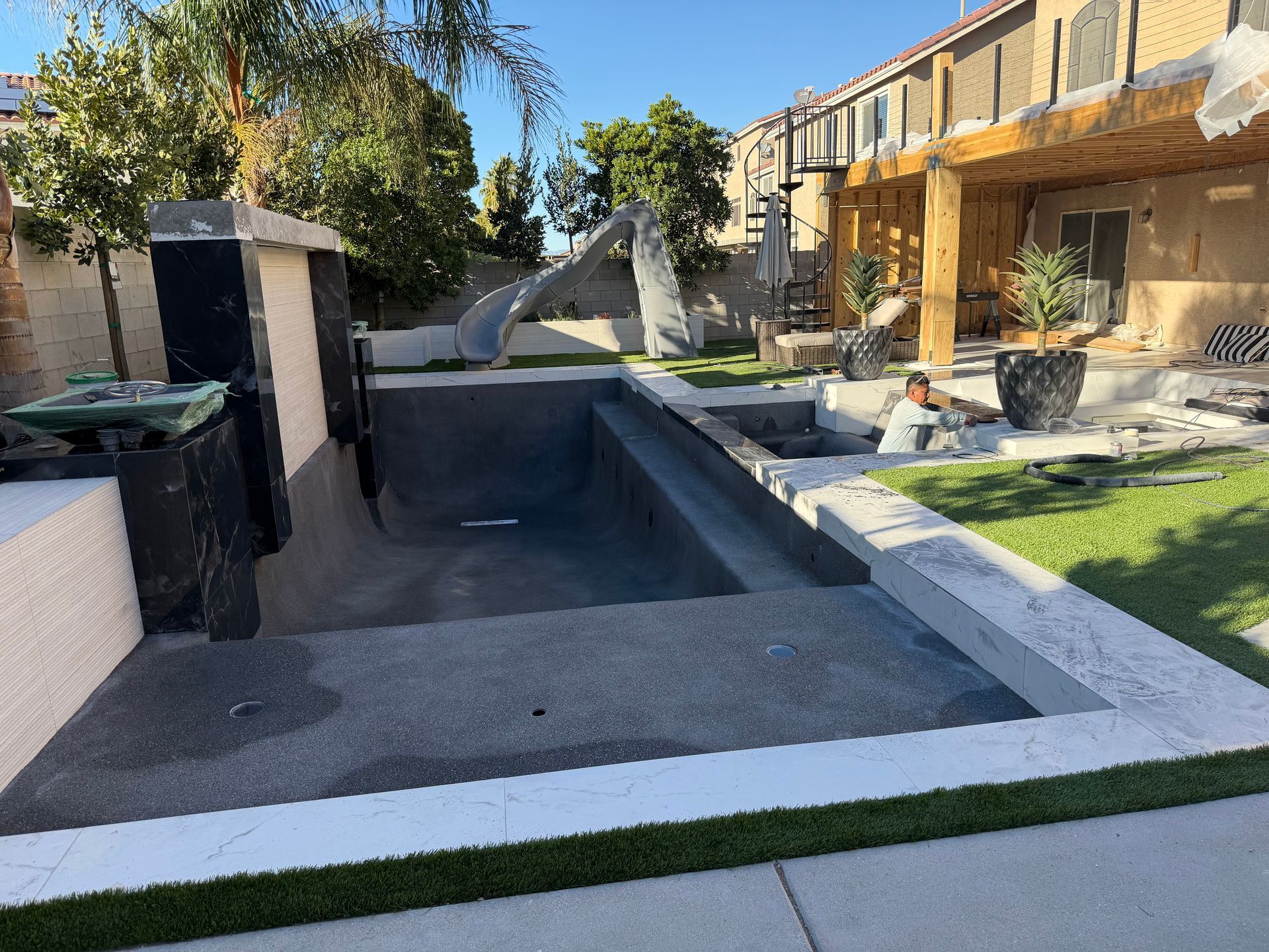 An empty concrete swimming pool under construction in a residential backyard with a worker visible in the background.