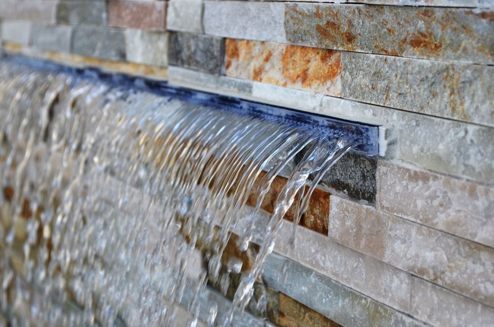A stone tile wall features a built-in water feature with clear water cascading down in a thin, continuous sheet.