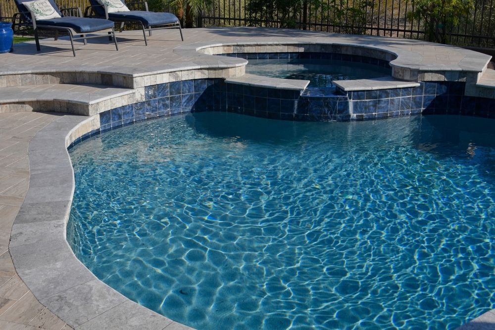 A backyard swimming pool with a stone patio, lounge chairs, and a small raised spa featuring a waterfall spillway.