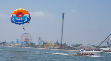 Parasailing over a waterfront amusement park with ferris wheels, a tall drop tower, and a speedboat below