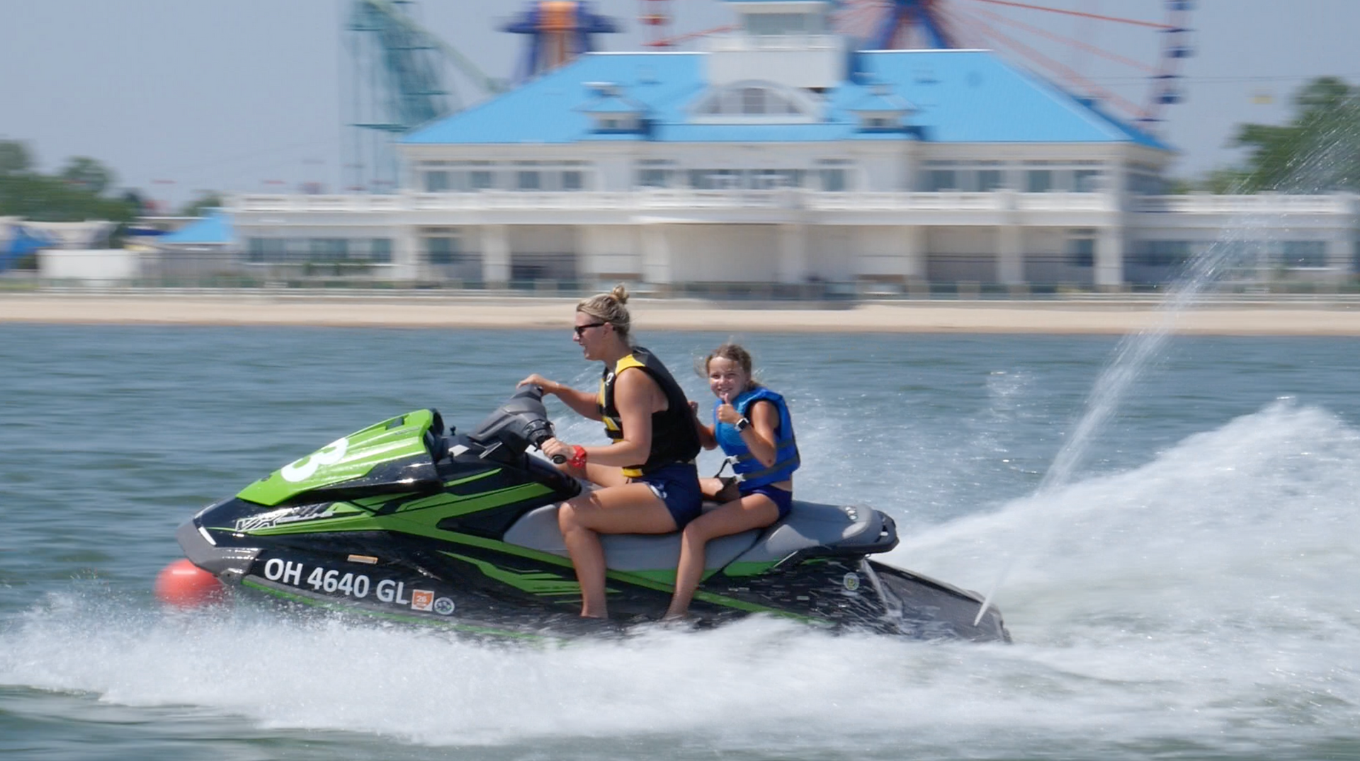 Two people riding a black-and-green jet ski, spraying water on a waterfront with a large building in the background.