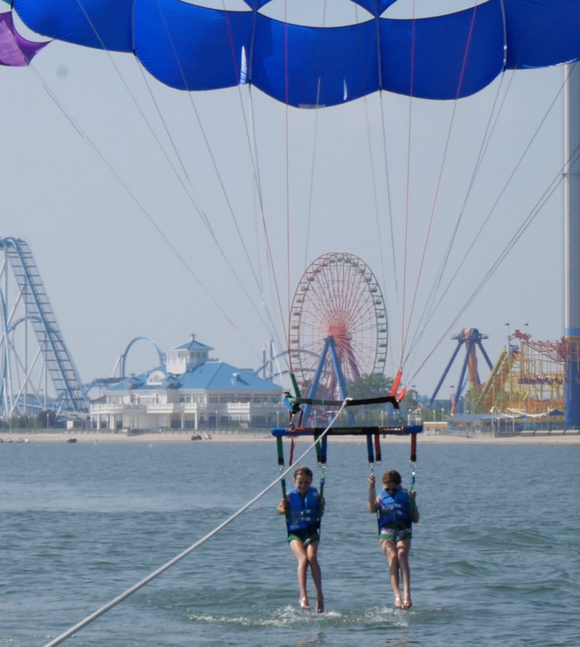 Two people parasailing over water with an amusement park and Ferris wheel in the background