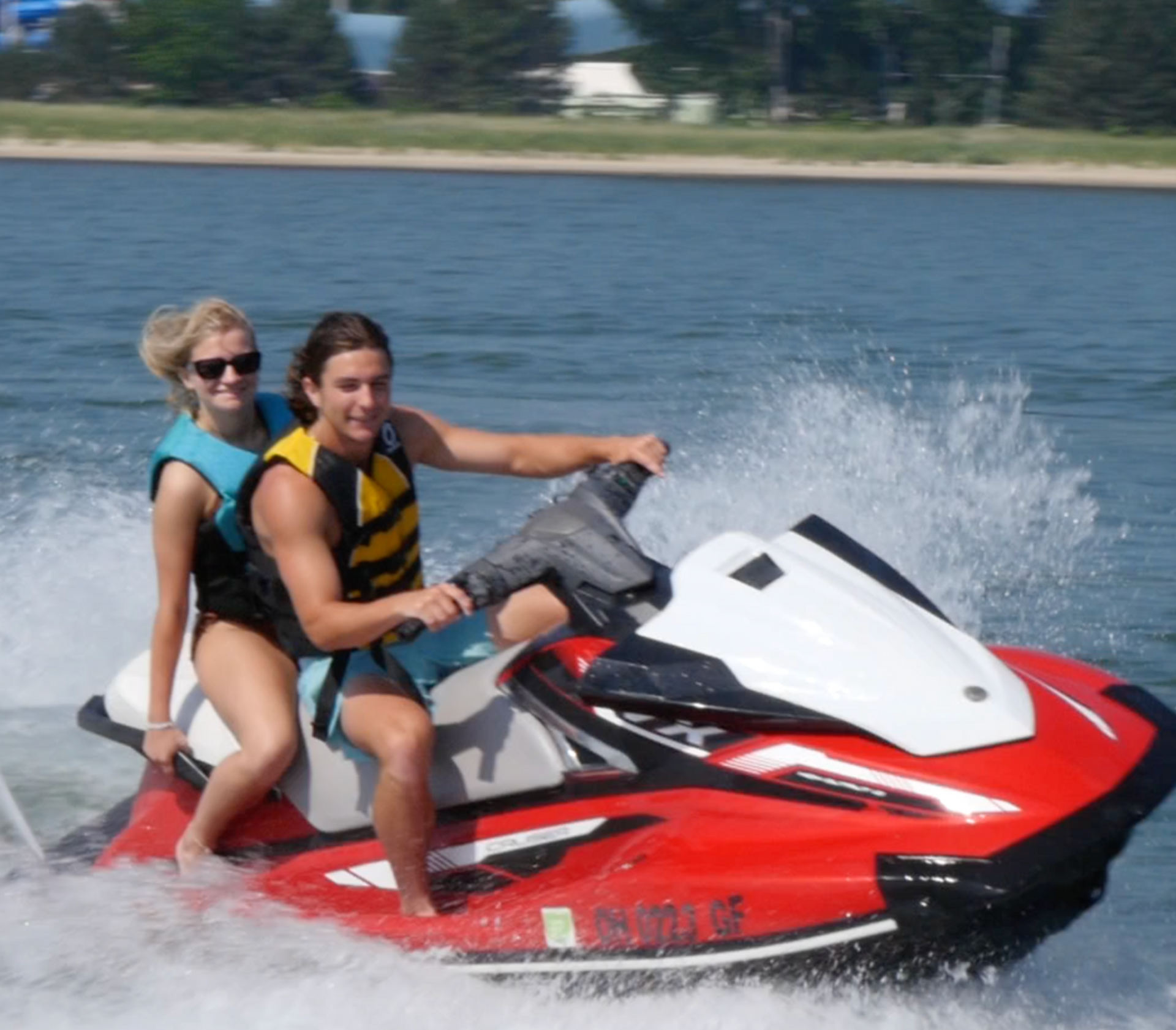 Two people riding a red and white jet ski on the water, spraying waves.