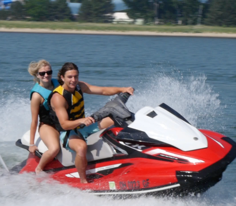 Two people riding a red and white jet ski on the water, spraying waves.