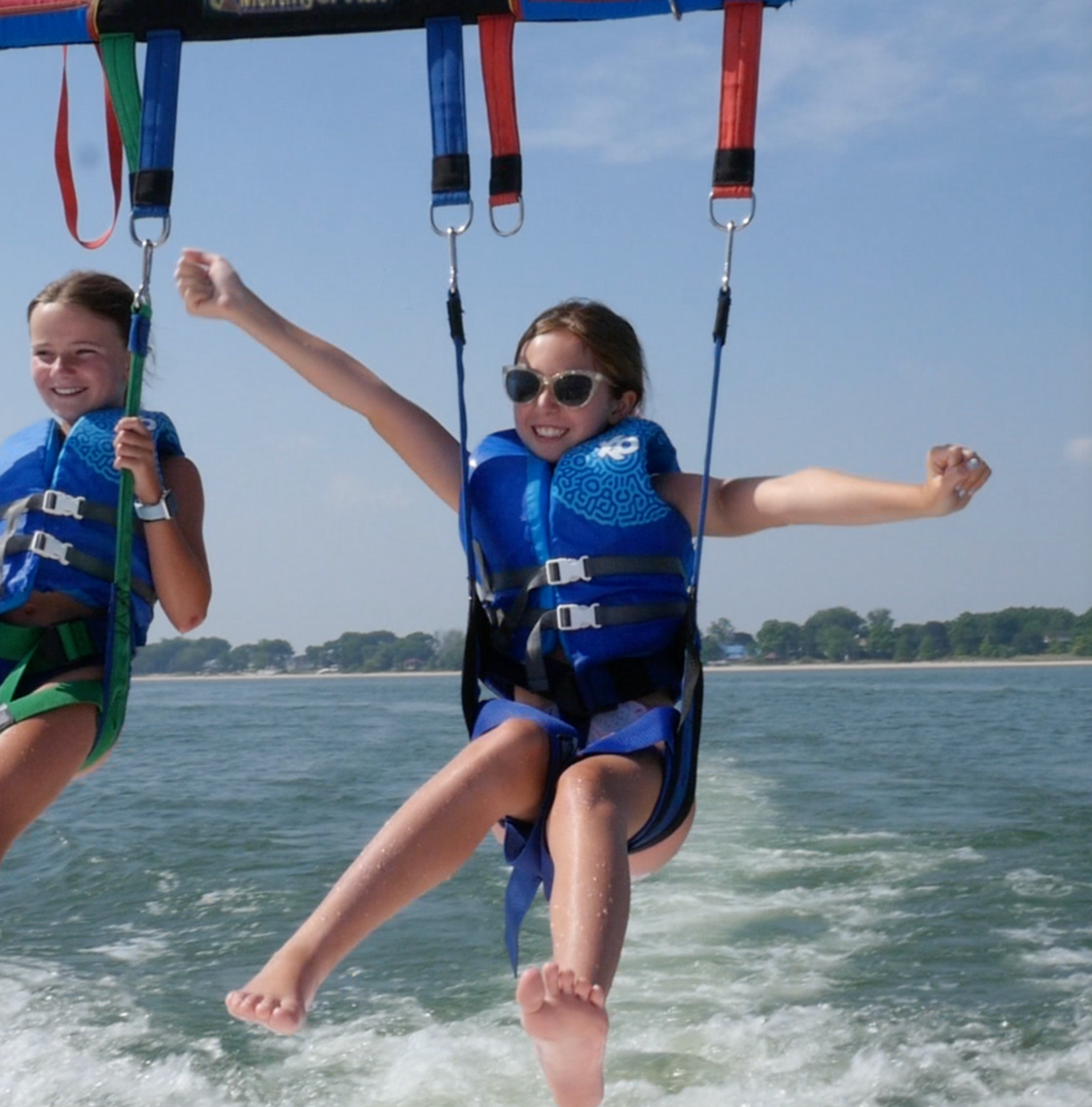 Two people parasailing over water, wearing blue harnesses and smiling against a bright sky
