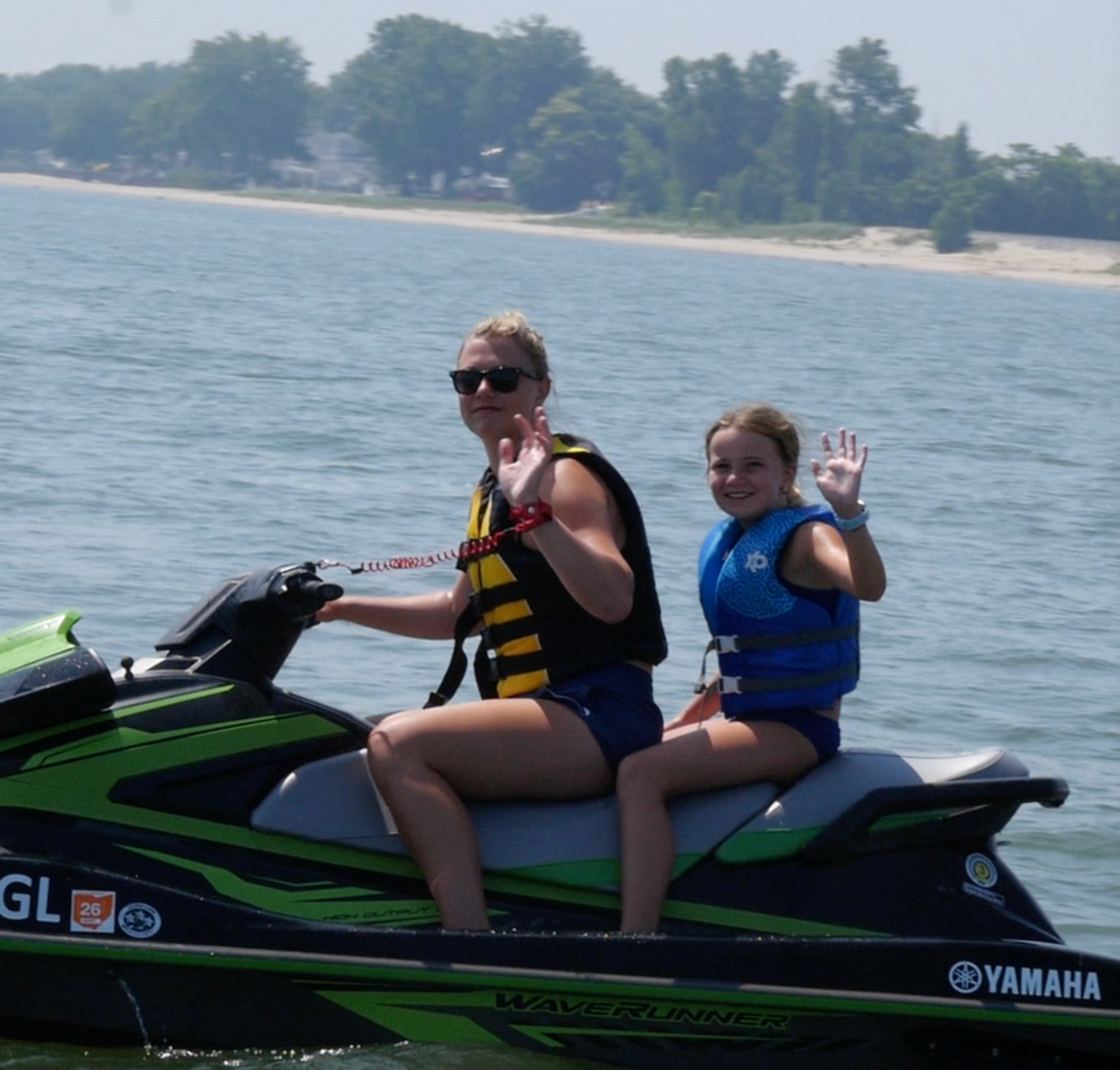 Two people waving while riding a green Yamaha jet ski on calm water near a sandy shore