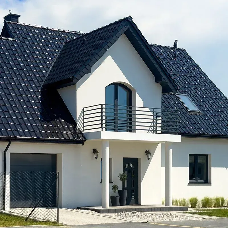 A modern white house with a black-tiled roof, a garage, a front balcony, and a dark entrance door under a clear blue sky.