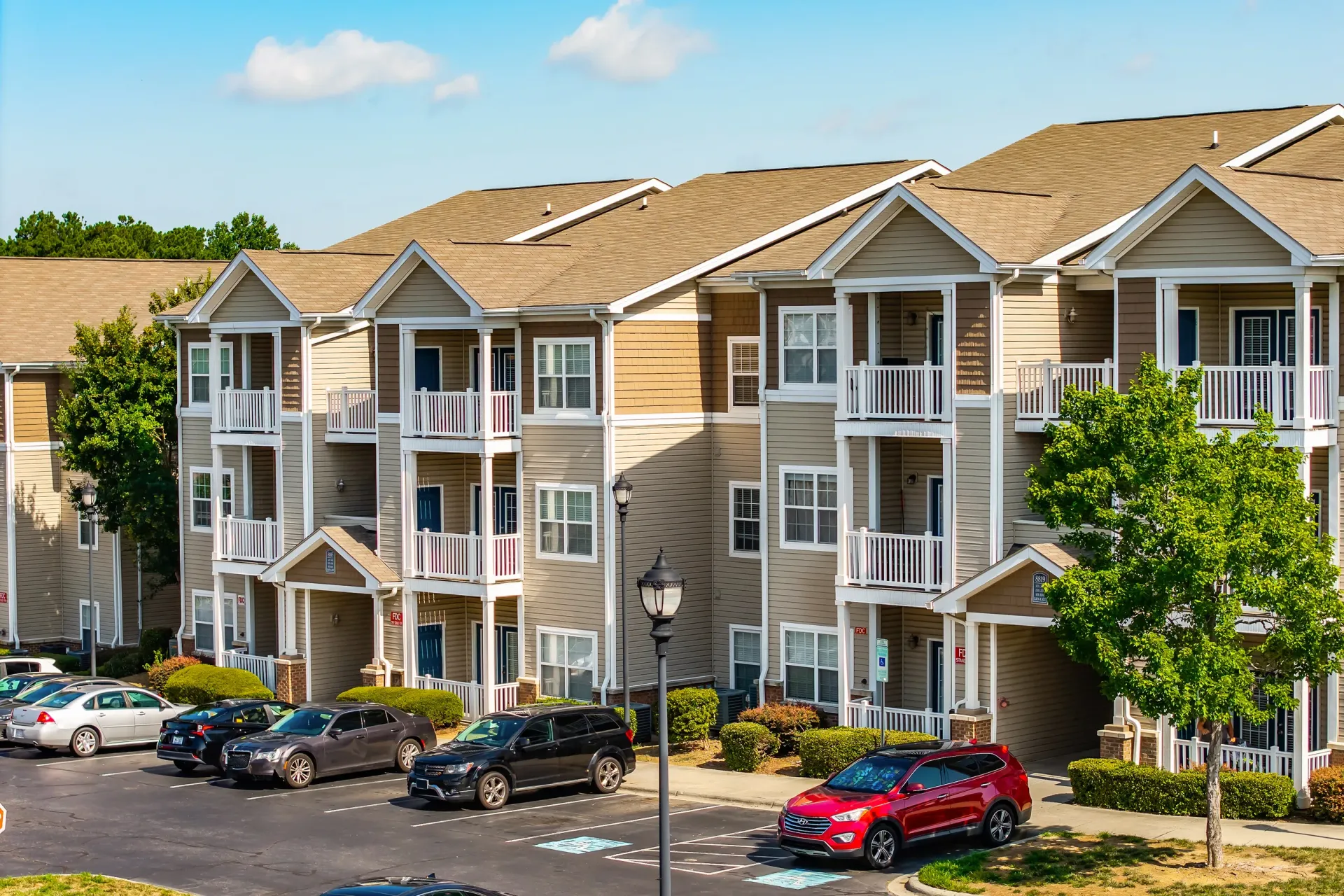 Exterior of a multi-story apartment building with balconies and a parking lot in front.
