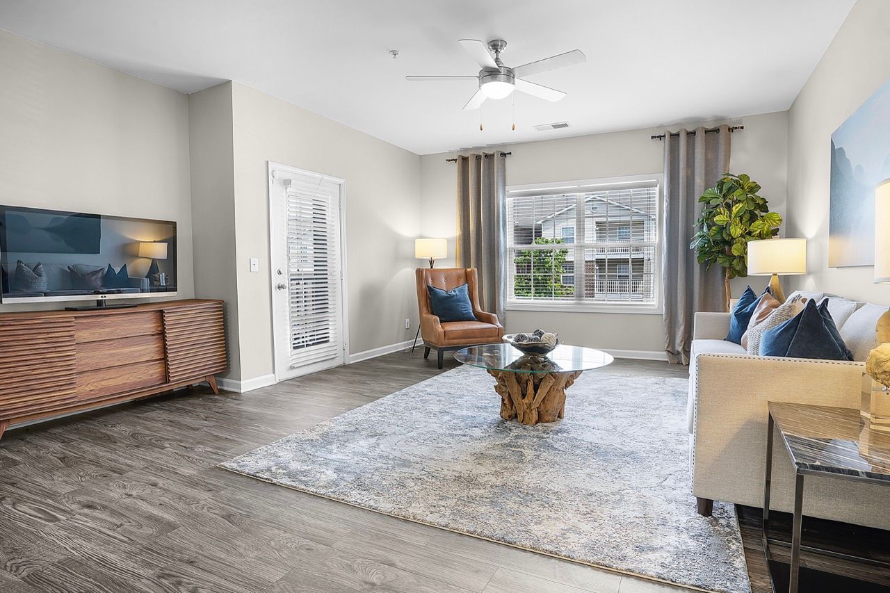 Living room with a sofa, armchair, glass coffee table, TV stand, and large window with curtains at Reserve at Stone Hollow in Charlotte, NC.