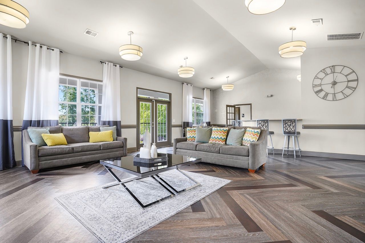 Bright apartment community lounge with two gray couches, a glass coffee table, and a large clock on the wall at Reserve at Stone Hollow in Charlotte, NC.