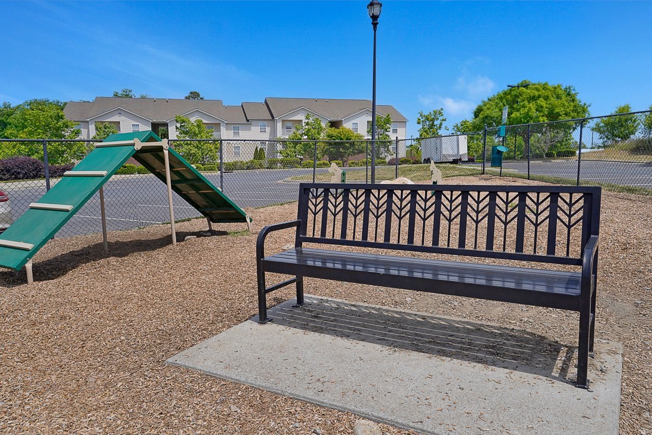 Metal bench on a concrete pad beside a green playground, with apartments in the background at Reserve at Stone Hollow in Charlotte, NC.