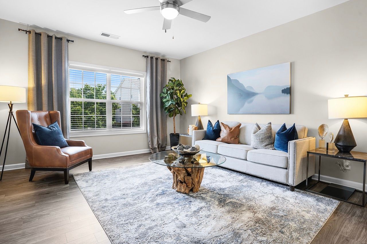 Bright living room with gray sofa, brown armchair, glass coffee table, and large window with curtains at Reserve at Stone Hollow in Charlotte, NC.