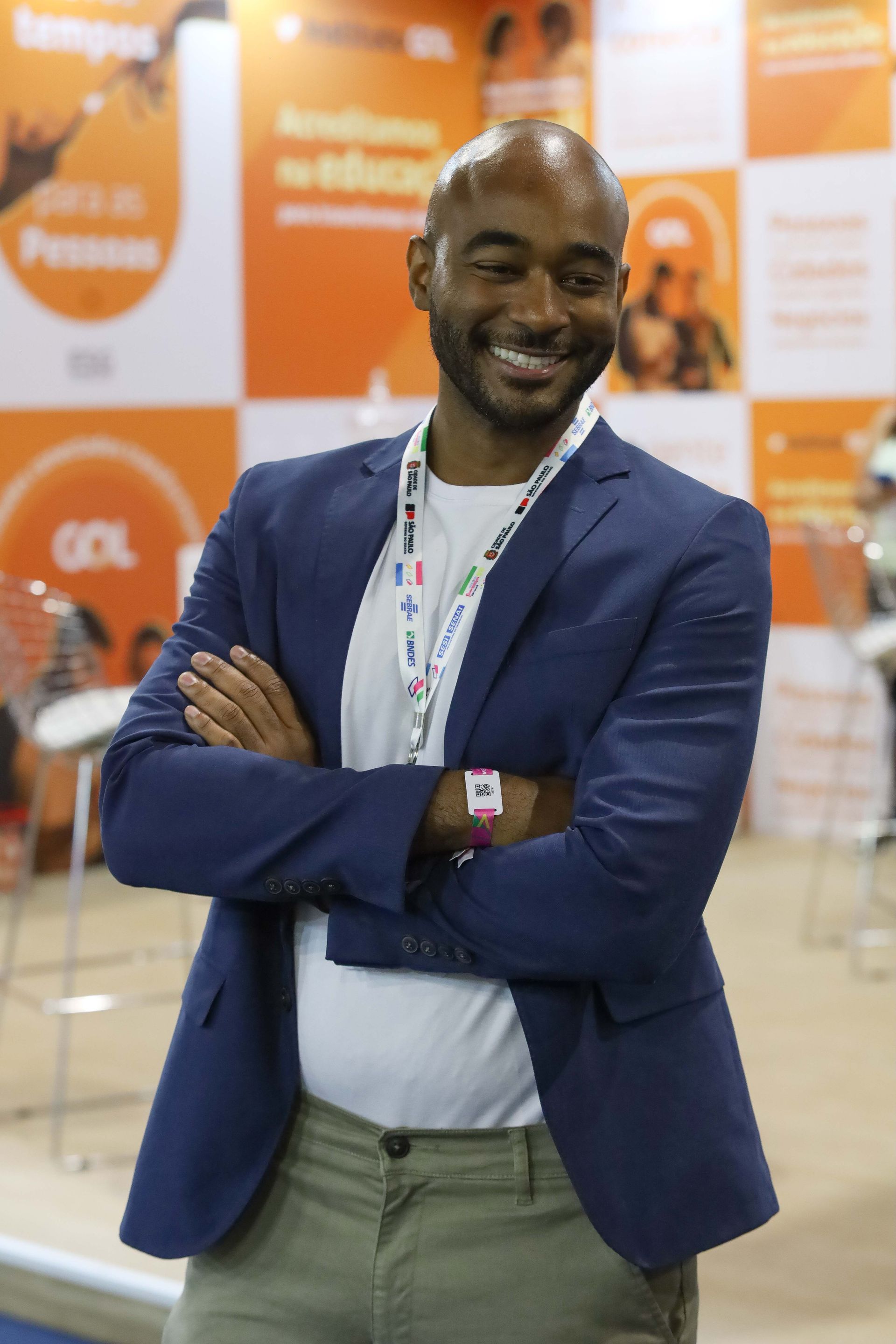 Um homem sorridente, vestindo um blazer azul, está de braços cruzados em um estande de eventos laranja e branco.
