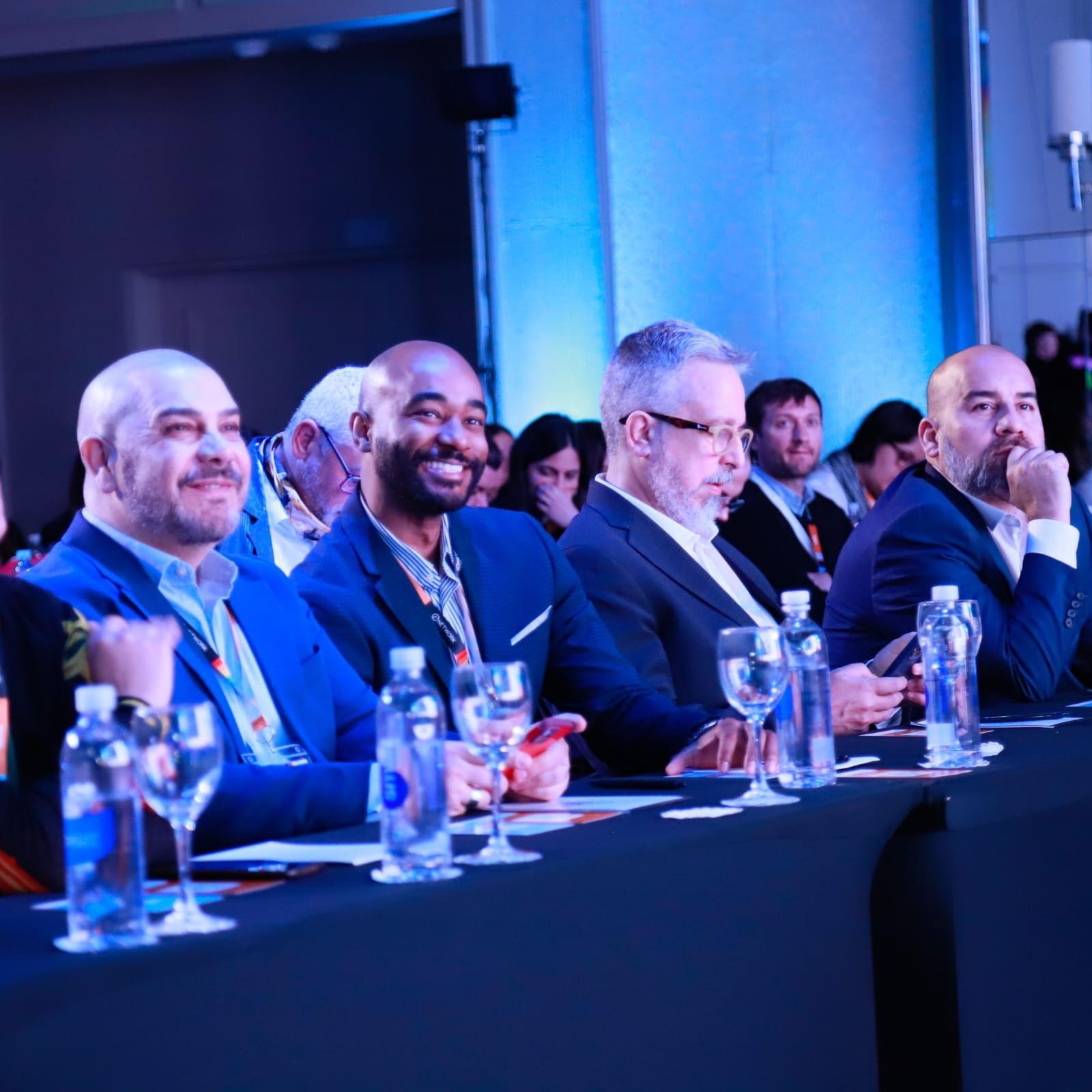 Membros da plateia sentados em uma mesa de conferência sob iluminação azul, sorrindo e assistindo ao evento.