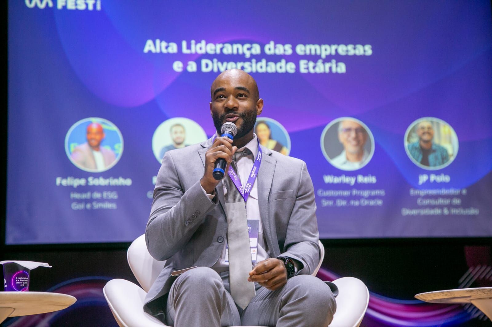 Palestrante no palco de uma conferência sobre diversidade, sentada com um microfone em frente a uma tela de apresentação azul-arroxeada.