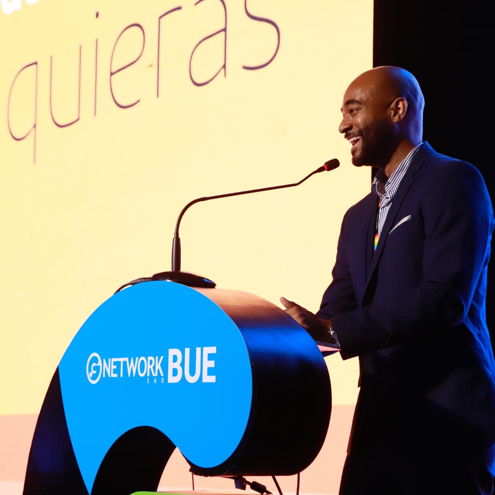 Homem discursando em um pódio azul da Network Blue no palco, com um fundo amarelo e um microfone.