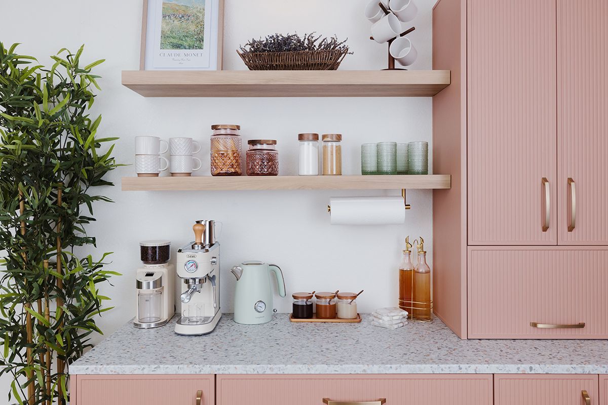 Modern kitchen shelf with coffee makers, jars, cups, and pink cabinet beside a leafy plant