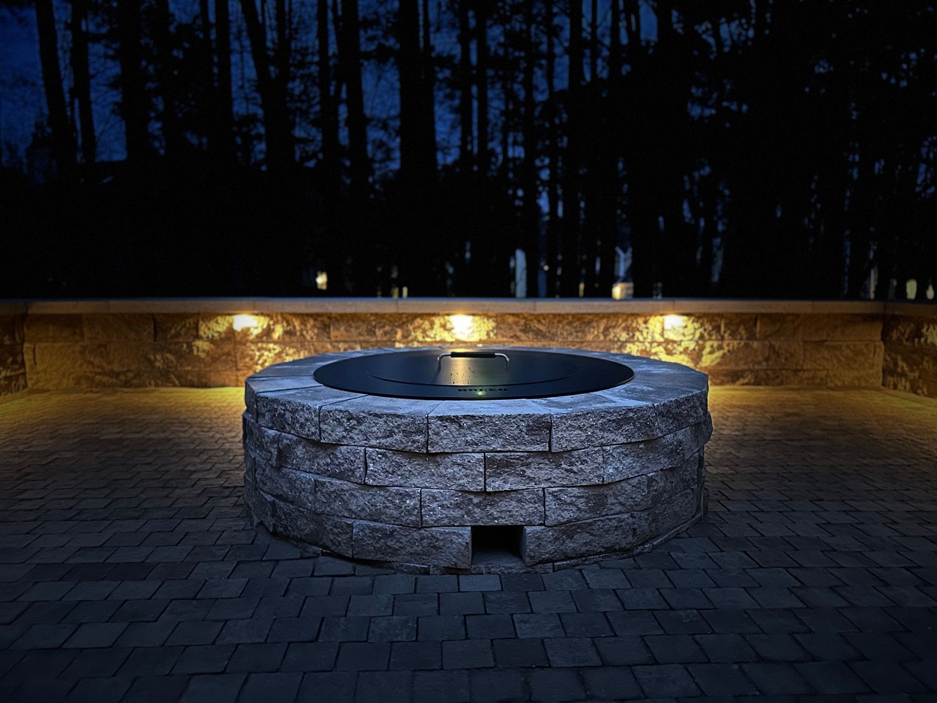 Circular stone fire pit illuminated at night, built into a paved patio with surrounding wall lights, forest in background.