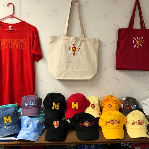 Red and cream merchandise display with T-shirts, tote bags, and embroidered caps on a wall shelf