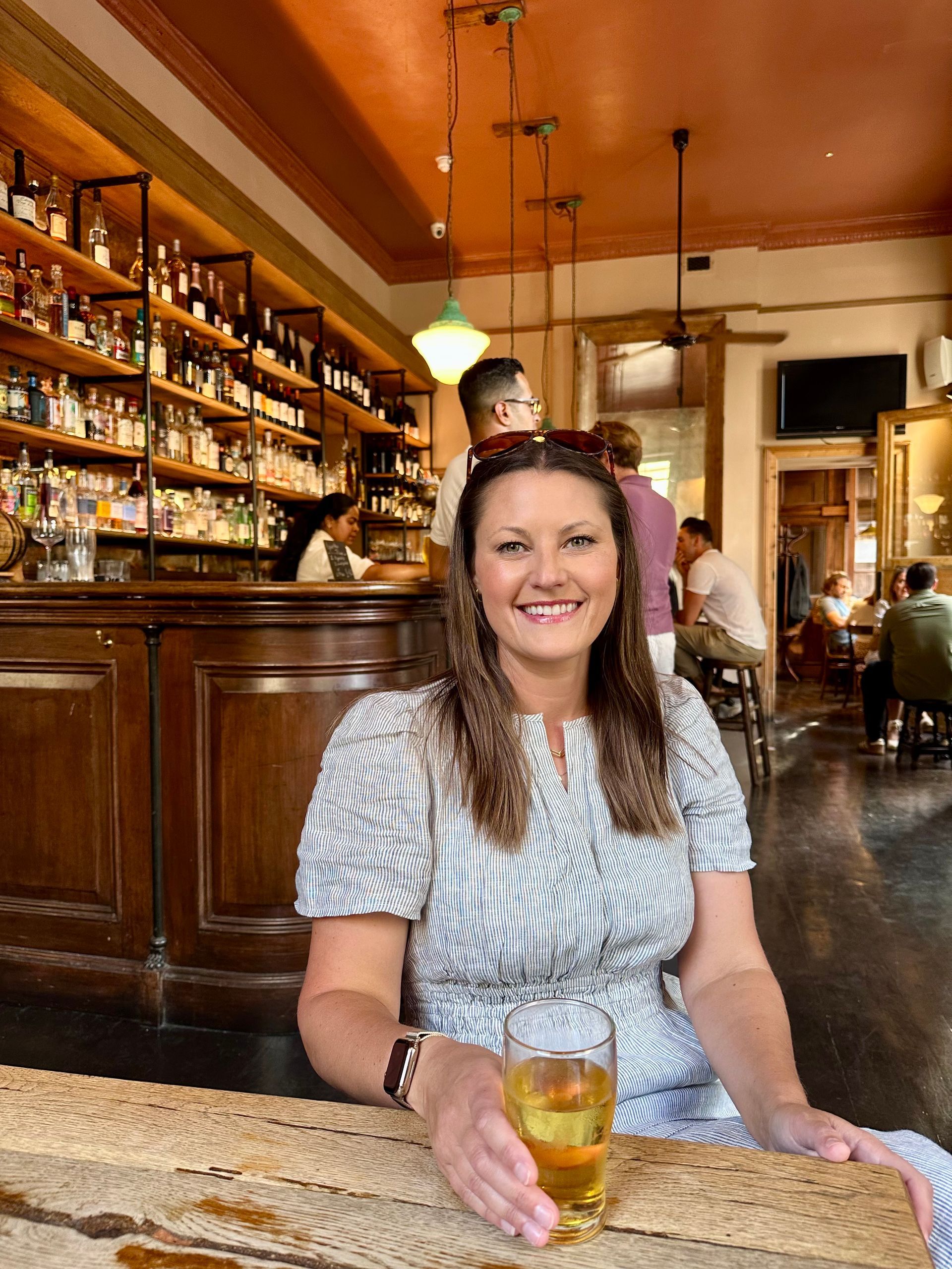 Woman smiling with a drink at a cozy bar, seated near shelves of bottles and warm wood decor.