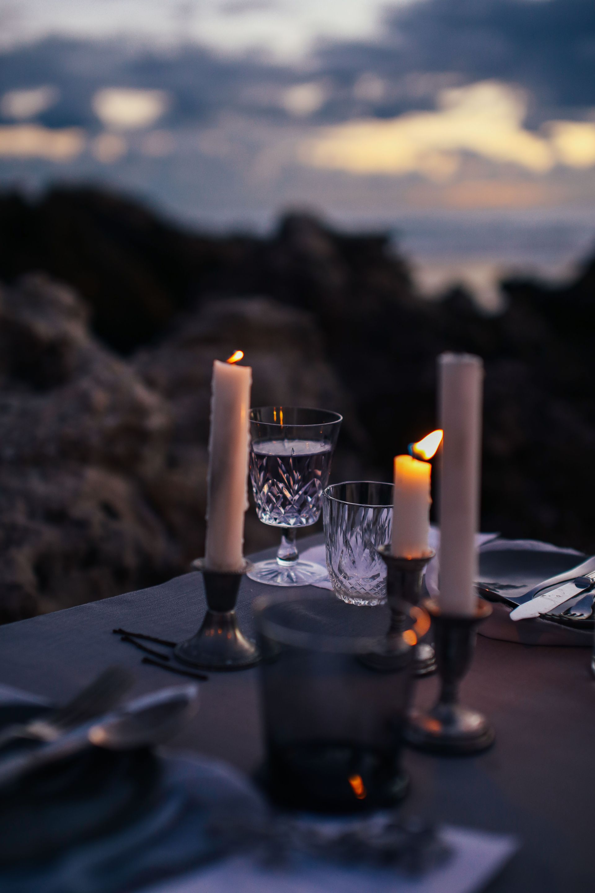 Candlelit dinner table by the sea at dusk, with wine glasses and blurred sunset background