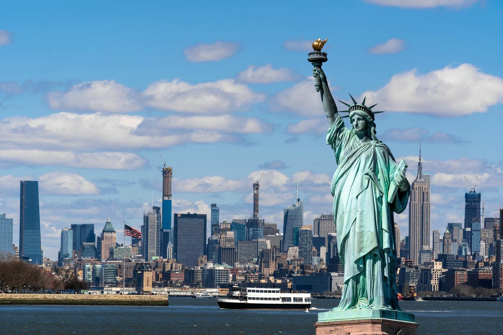 The statue of liberty is in front of a city skyline with a boat in the water.