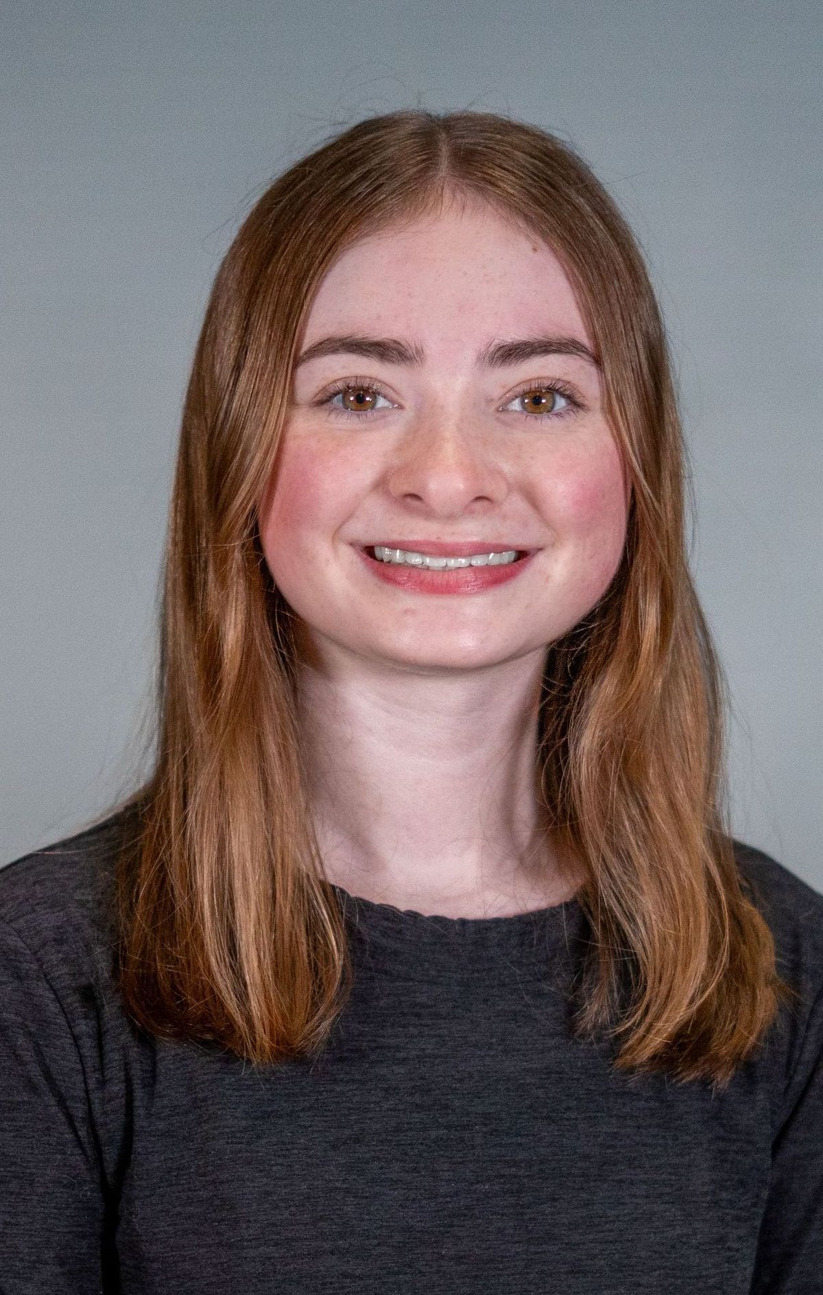 A smiling person with shoulder-length light brown hair wearing a dark grey sweater against a plain grey background.