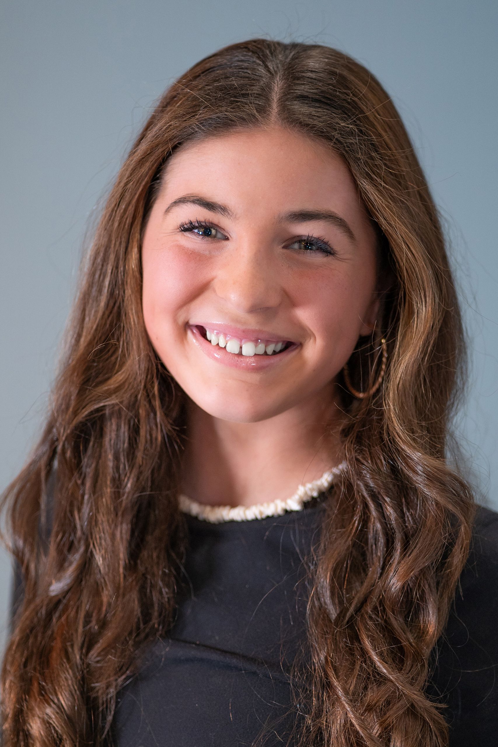 A smiling young person with long brown hair and a beaded necklace against a plain blue background.