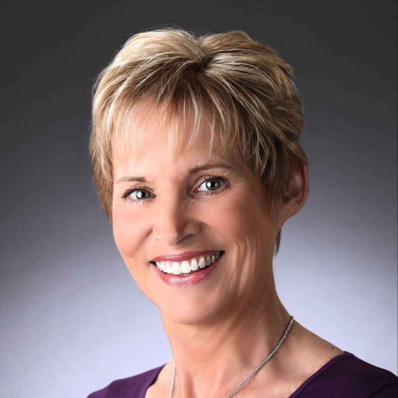 Woman with short blonde hair, smiling at camera; wearing a purple top and silver necklace.