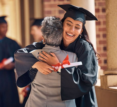 Graduate in cap and gown embraces another person holding diploma, smiling outside a building.