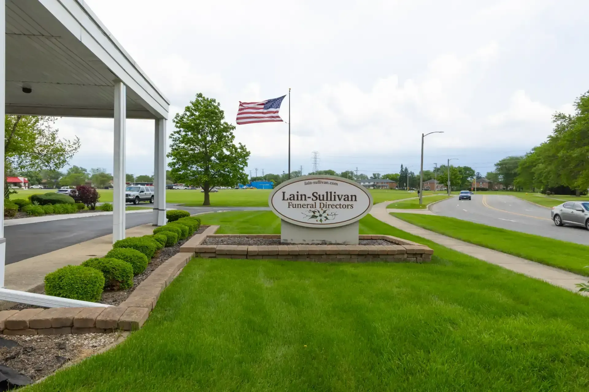 Lia-Sullivan sign beside an American flag and green lawn at a roadside entrance