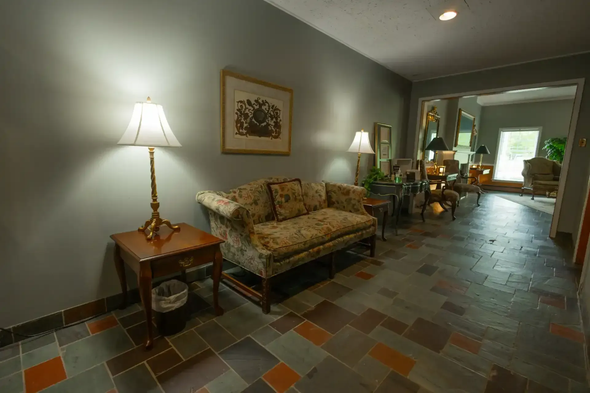 Dim living room with patterned sofa, lamp, tiled floor, and hallway leading to a bright room.