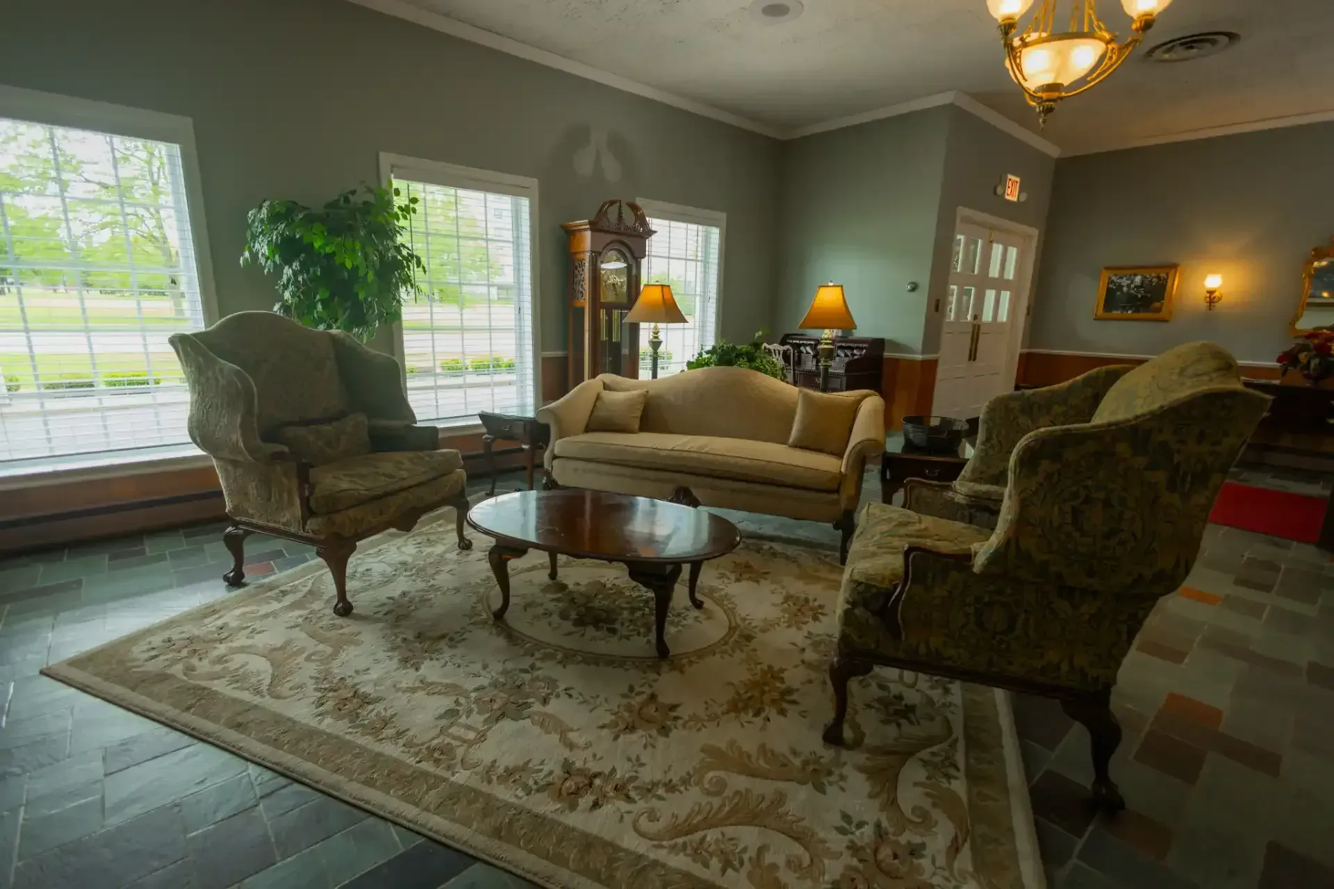 Traditional living room with green walls, floral rug, armchairs, sofa, coffee table, and large windows.