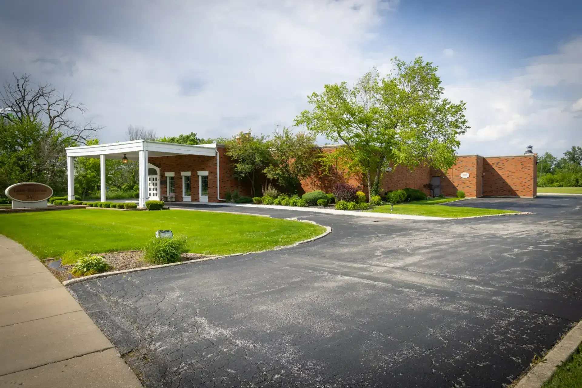 Brick building with white entrance canopy, green lawn, and empty asphalt driveway under cloudy sky