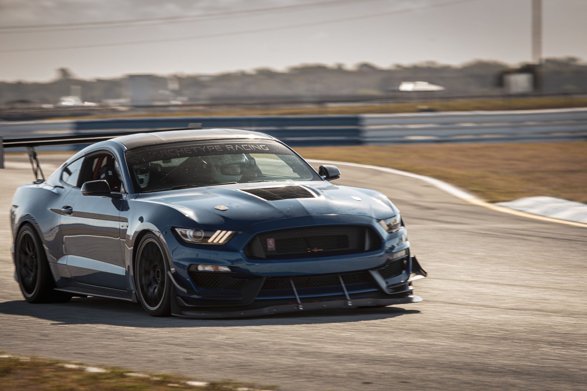 Blue Ford Mustang race car on a track, with a large spoiler and aggressive bodywork. Sunny day.