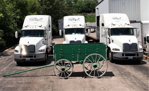 Three white semi trucks parked behind a green wagon in a lot beside a building.