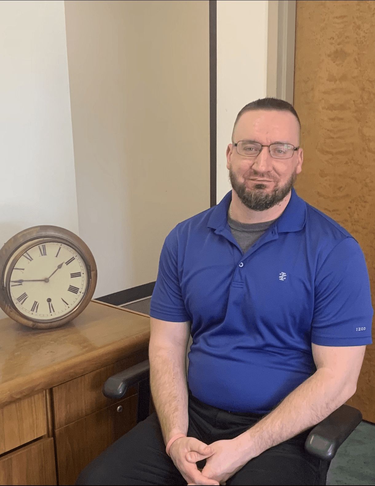 Man seated at a desk beside a large round clock in a simple office setting.