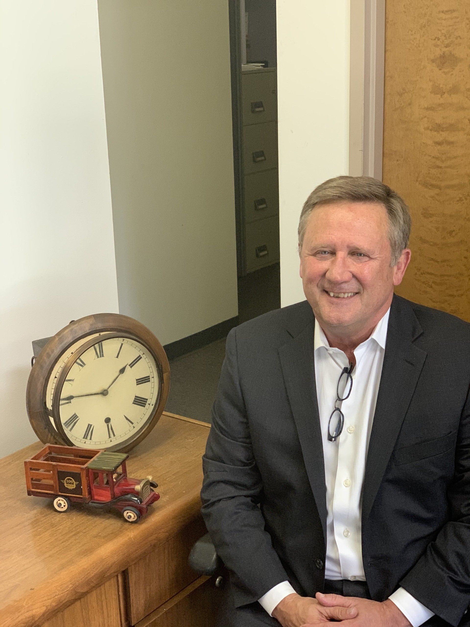 Smiling man in a dark suit seated beside a clock and toy truck on a wooden table.