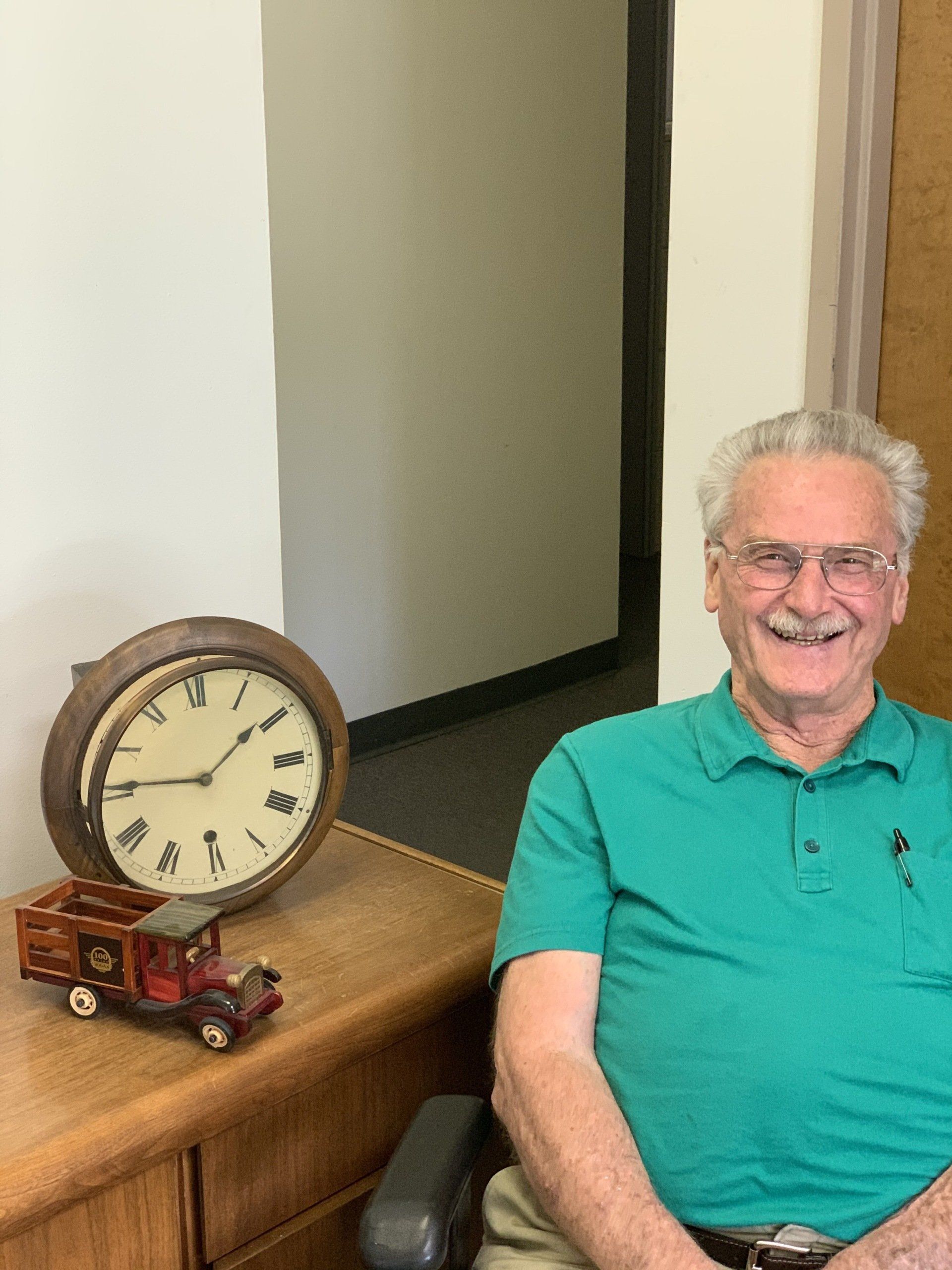 Smiling man in green polo sitting beside a table with a vintage clock and toy truck