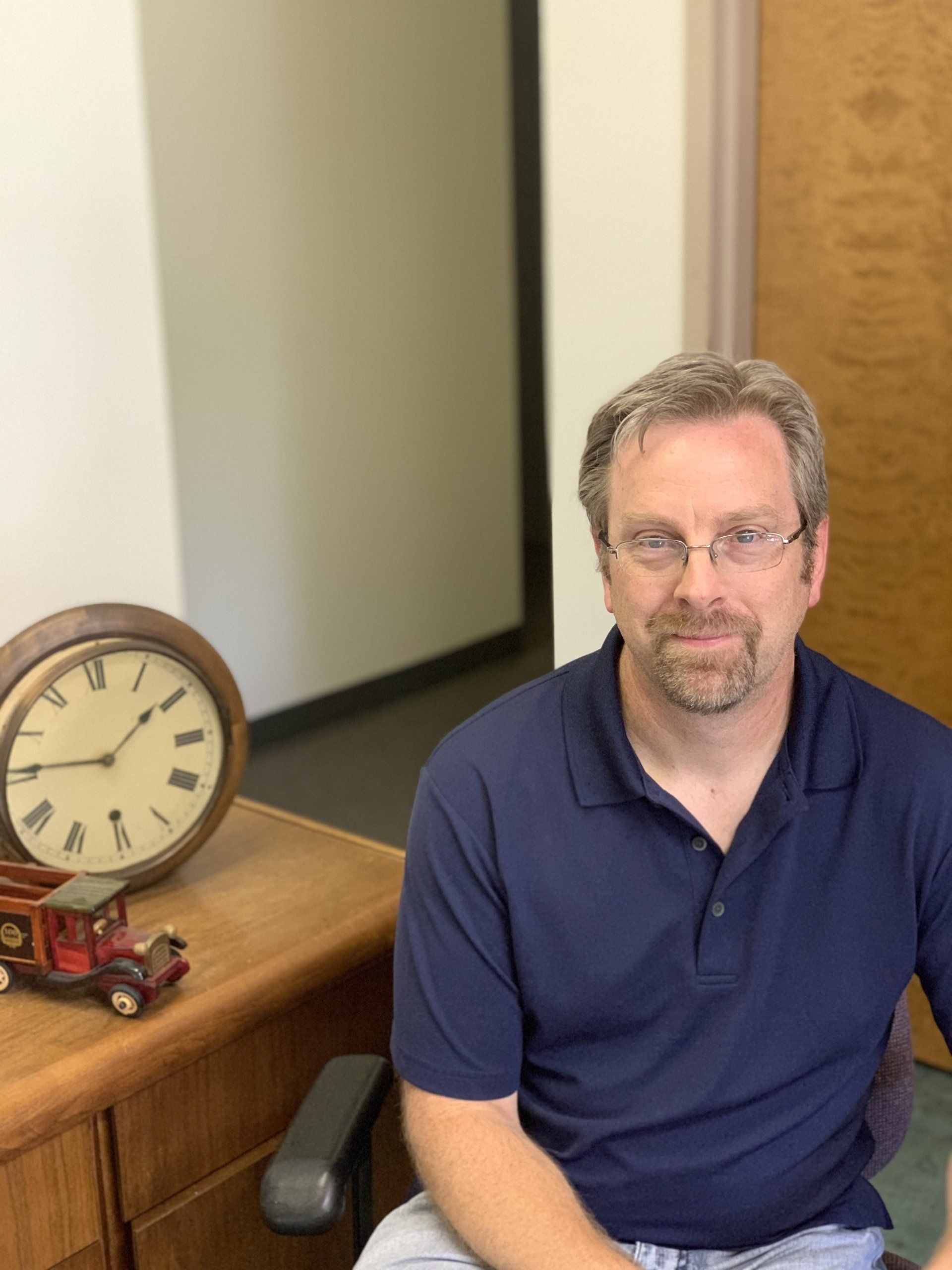 Man seated at a desk beside a clock and toy truck, wearing a navy polo shirt and glasses.