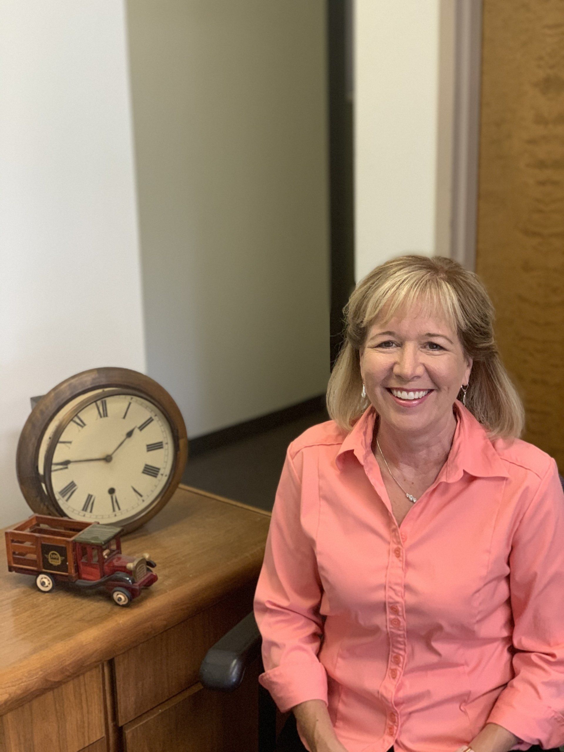 Woman smiling beside a wooden table with a clock and red toy truck in an office setting