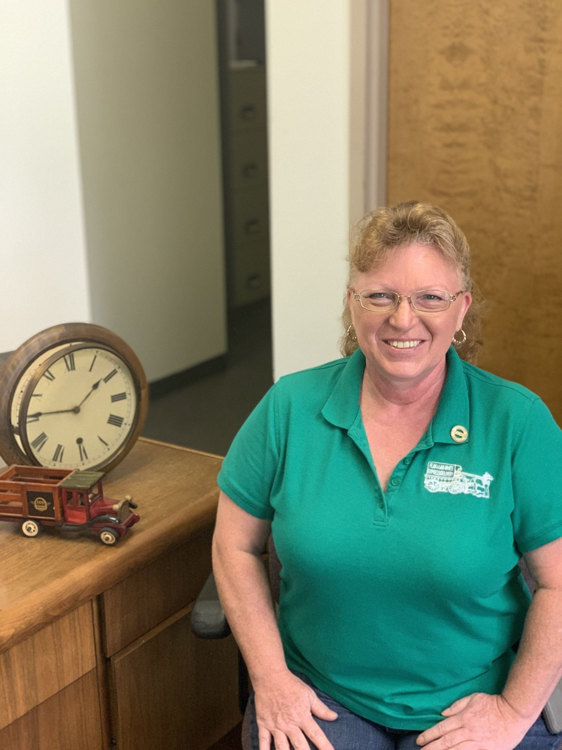 Woman in green polo smiling beside a wooden clock on a counter in an office hallway