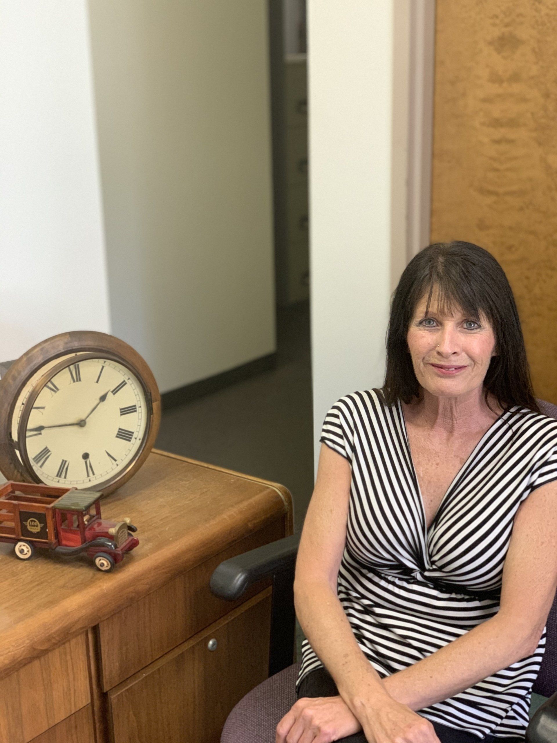 Woman seated beside a wooden desk with a clock and small red toy car in an office hallway.