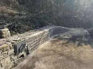 Stone retaining wall along a dirt path in a wooded outdoor area with a small plaque on the left