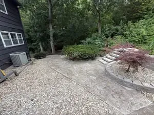 Gravel patio beside a dark house, with stone steps, curved retaining wall, and trees in the background