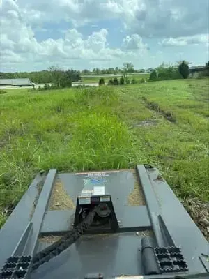 View from a mower driving through tall green grass under a cloudy sky, with fields and buildings ahead