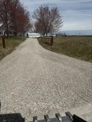 Gravel driveway leading to a house, flanked by leafless trees and open fields under a cloudy sky