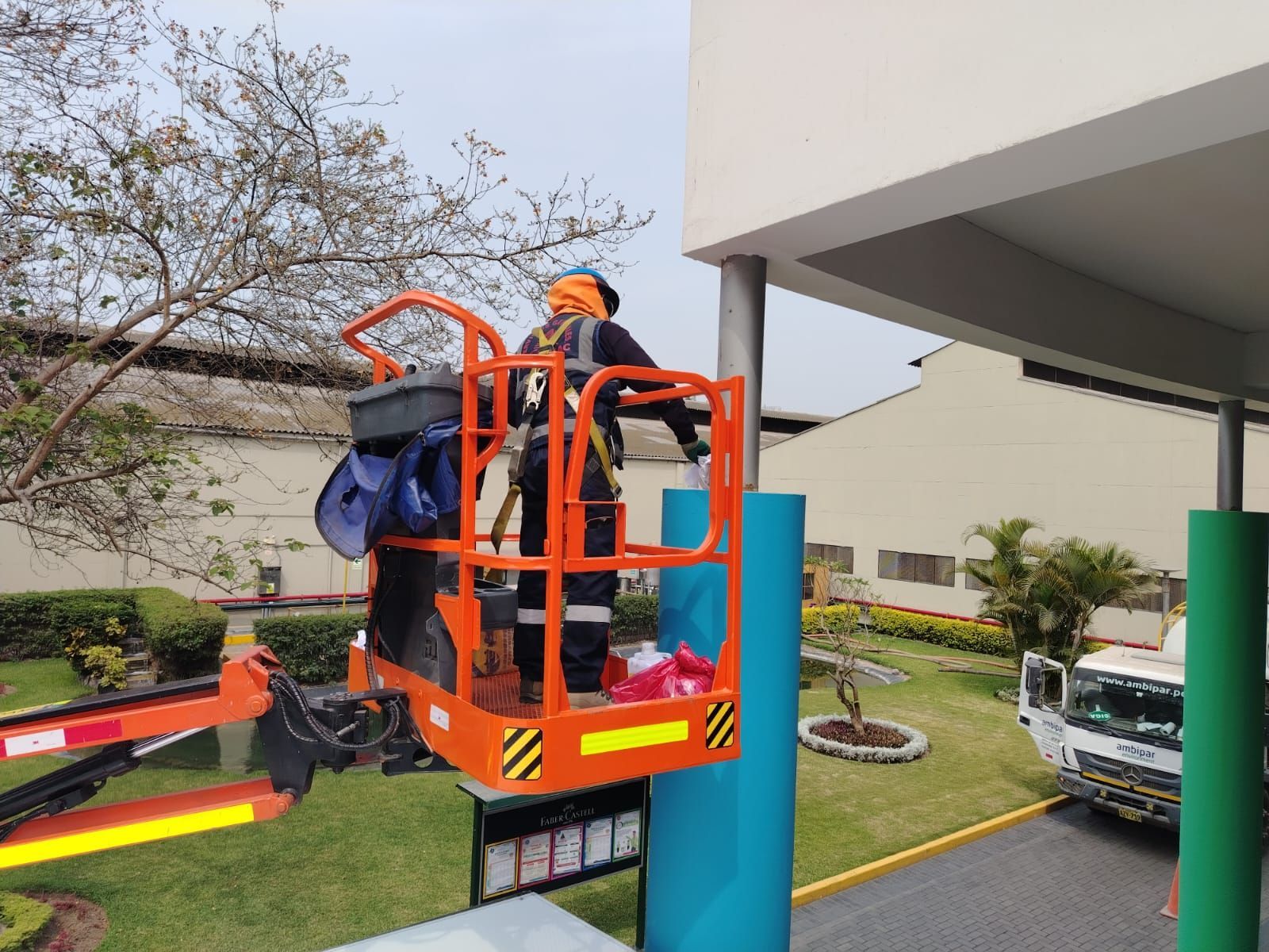 Persona en un elevador naranja trabajando en un pilar azul afuera de un edificio; hay un camión estacionado cerca.
